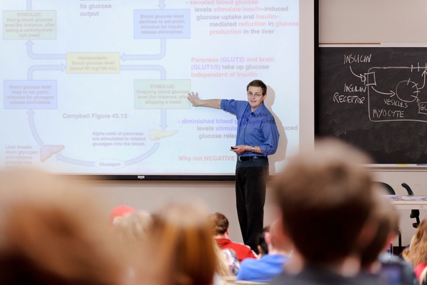 David Abbott stands at the front of a classroom full of students, gesturing to a slide on a projector screen. Photo credit: UW-Madison
